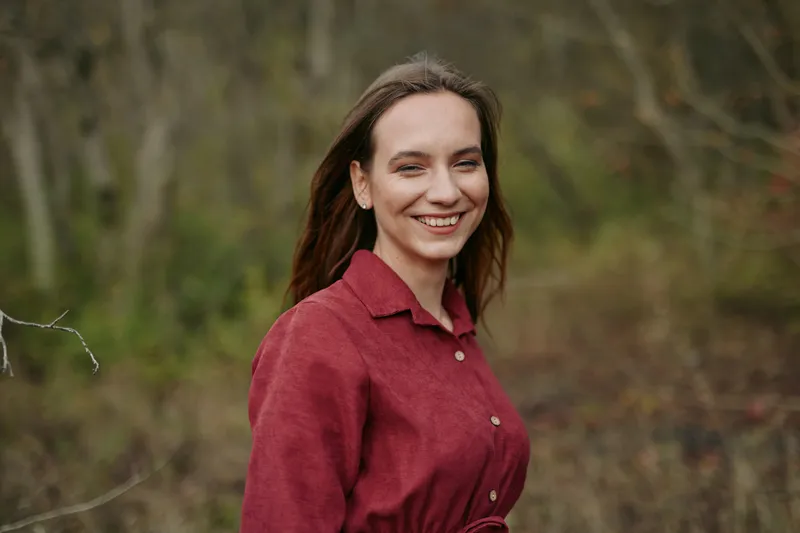 A smiling woman in a red dress stands in woodland, captured with natural light and calm posture, offering a warm, genuine portrait that shows authenticity and credibility.