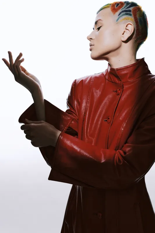 Stylish woman with colorful shaved hair poses gracefully in red leather coat, showing elegance, confidence, and modern fashion in a clean white background studio setting.