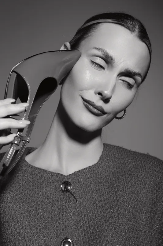 Elegant Caucasian woman with fair skin holds high heel shoe close to face, eyes closed, wearing textured sweater in black and white portrait.