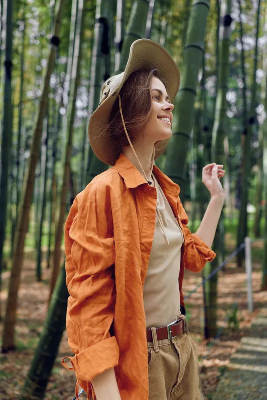 Woman bamboo forest hat orange portrait of a smiling young female in casual jacket and boho outfit, walking through serene greenery with sunlight filtering for travel and nature mood.