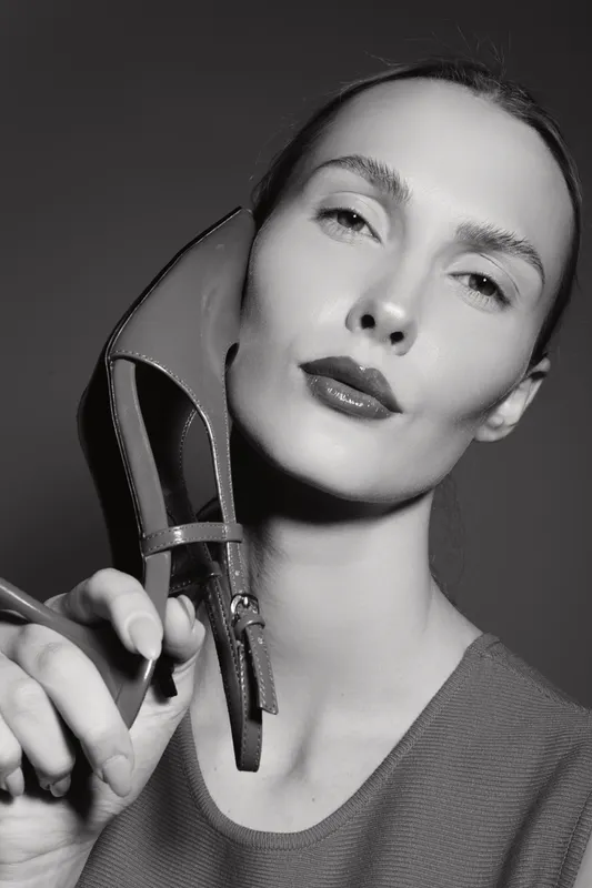 Close-up of a fair skinned white woman holding a high heel shoe near her face in black and white studio portrait.