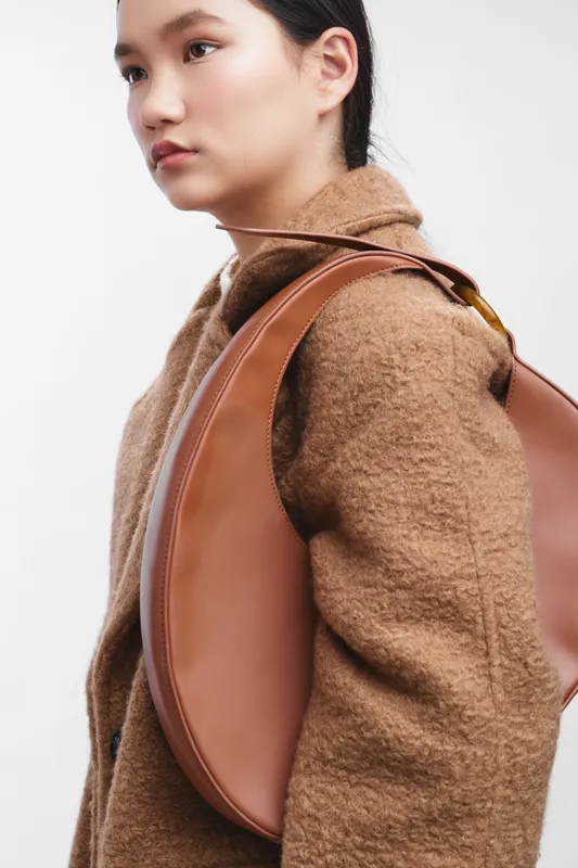 Asian woman wearing a tan coat with a leather bag over her shoulder stands poised in a clean studio, conveying confidence and style through minimalist fashion and contemporary grooming.