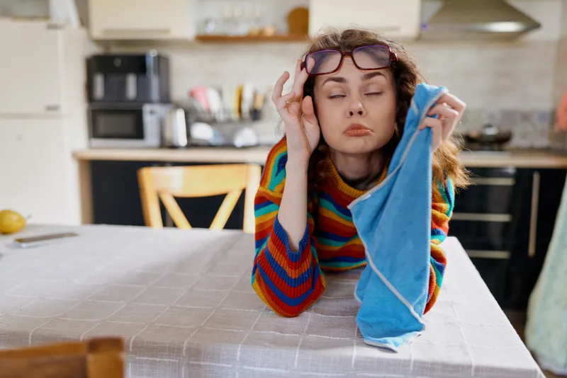 A tired woman in a striped sweater rests her head on her hand at a kitchen table, towel in hand, glasses on her head. Concept for fatigue and daily home life.