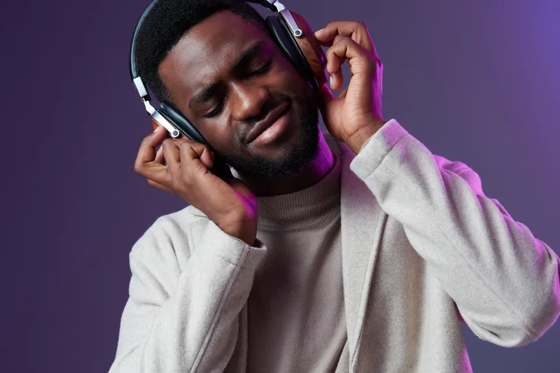 Young Black man enjoying music with headphones, showcasing a relaxed emotion against a vibrant purple background, perfect for themes of leisure and music appreciation