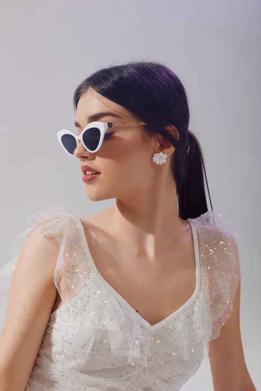 Young woman in white dress with sheer sleeves wears stylish sunglasses and floral earrings. She looks sideways with confident expression. Concept of woman beauty fashion and elegance.