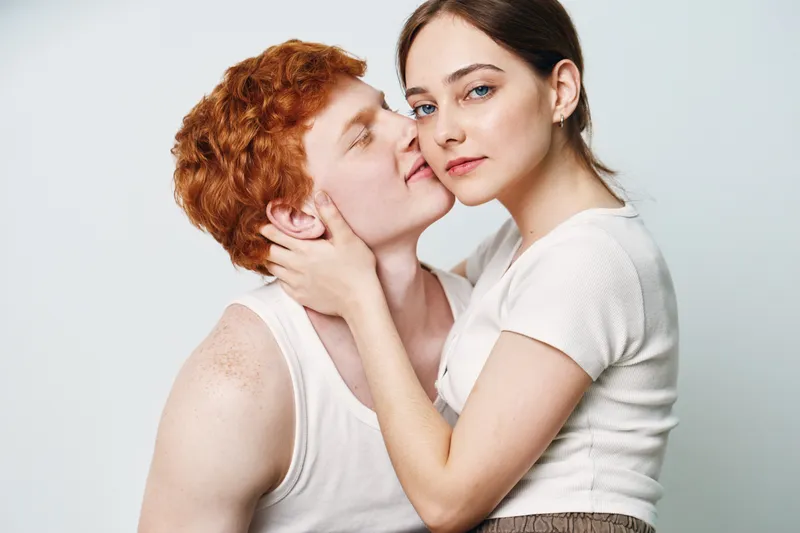 Couple with woman embracing man intimately with hands on neck, white casual clothes, studio portrait with light background, showing affection, connection, and closeness.