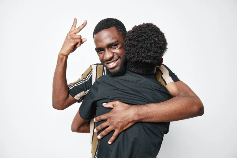 Two black men hugging and smiling with one showing peace sign in casual clothes on white background for friendship and happiness.