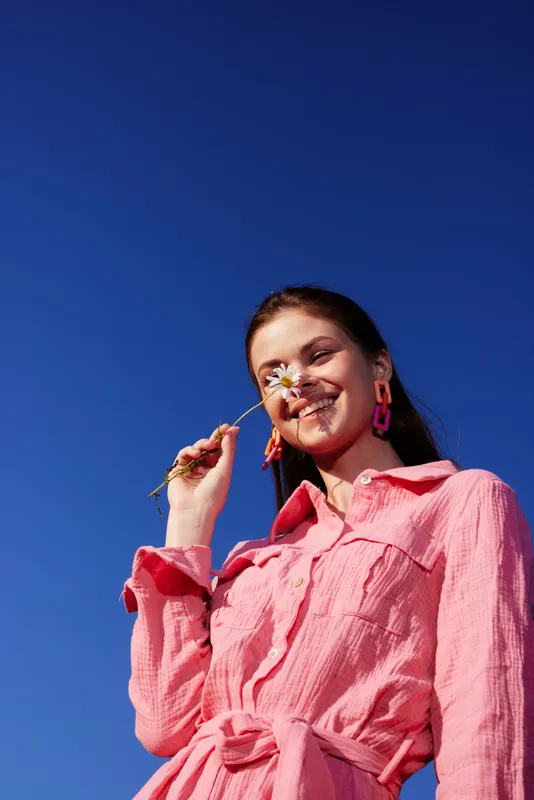 Smiling female model with flower against a blue sky, wearing a pink dress, showcasing joy and summer vibes, perfect for concepts like happiness and beauty