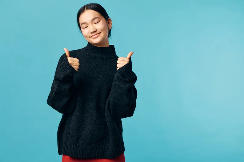 Young woman wearing black sweater shows thumbs up hand gesture with closed eyes on blue background. Positive expression, casual clothing and relaxed mood in studio wall shot.