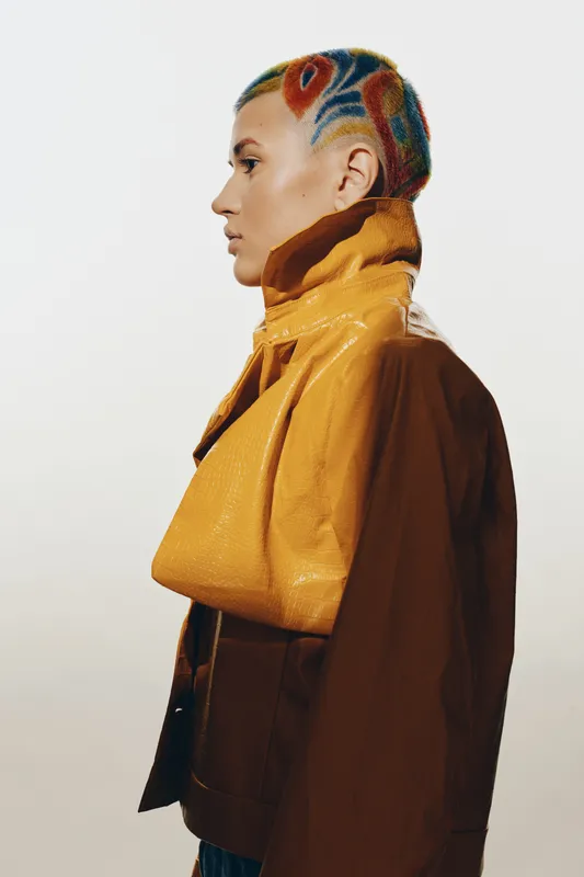 woman with colorful braided hair wearing a stylish brown and yellow jacket, side profile portrait with minimalist white background and modern fashion details