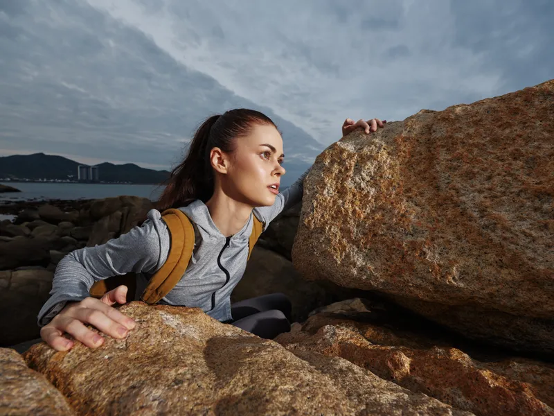 Young woman climbing rocky terrain, showcasing determination and resilience, wearing sports attire against a dramatic cloudy background