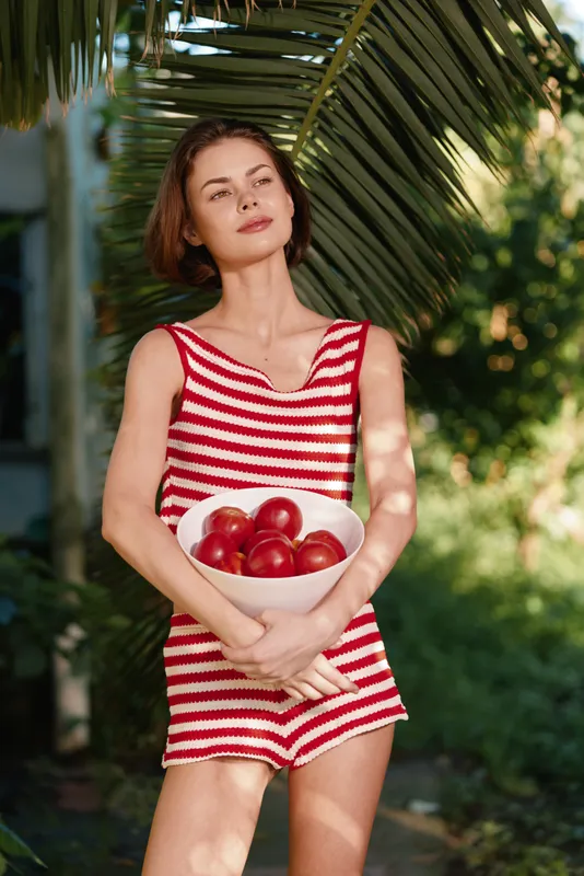 A young woman in a red and white striped swimsuit stands outdoors, holding a bowl of apples. Sunlit garden surroundings, lush greenery, and calm, relaxed mood create a summery, casual portrait.