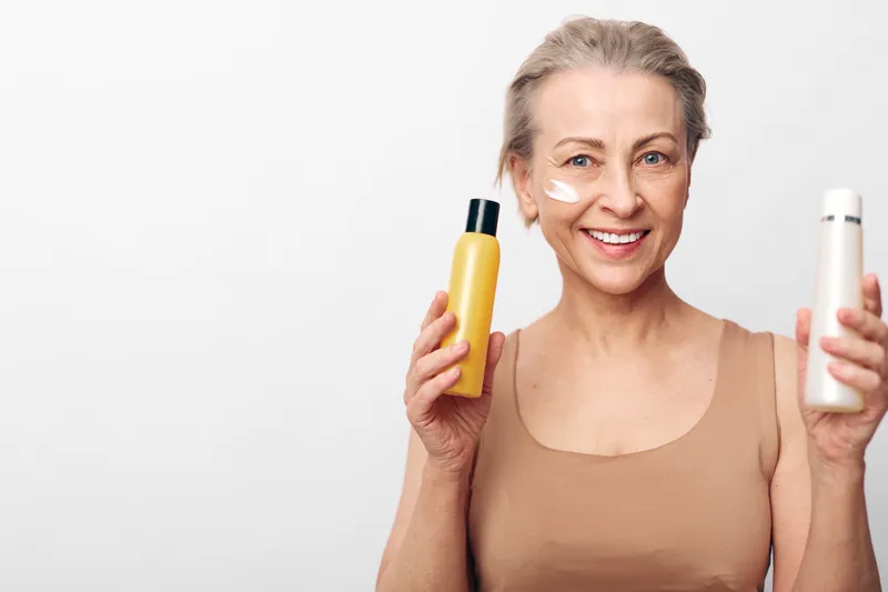 Smiling mature woman with short hair holding two cosmetic bottles, applying cream on cheek. Concept of skincare, beauty routine, and healthy aging with natural products on white background.