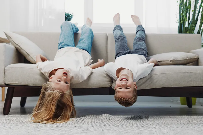 Two happy children lying upside down on a beige sofa in bright living room, smiling and having fun. Concept of childhood joy, playfulness, and family time at home.