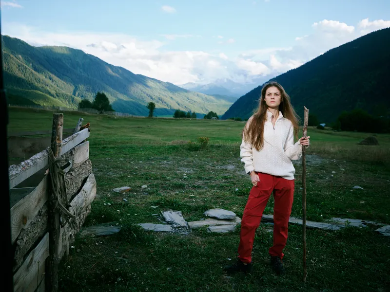 Girl standing with a wooden stick in a rural meadow, mountains in the distance and a rustic fence, nature scene capturing outdoor adventure and countryside beauty