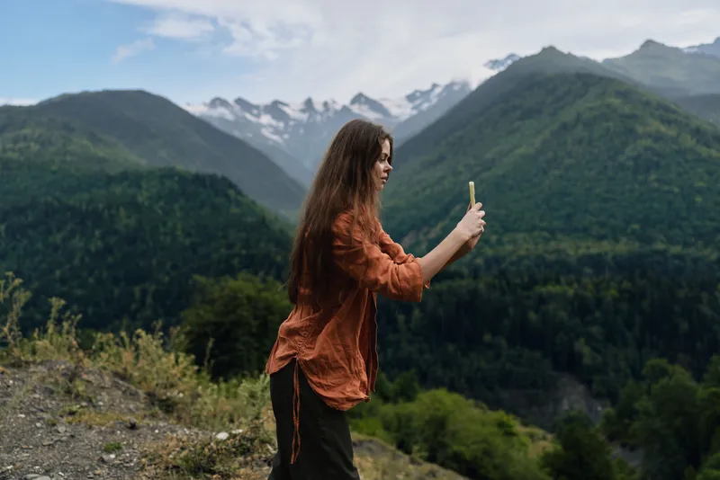 Woman on a hillside in a mountainous landscape, taking a photo with a smartphone, capturing expansive scenery and the beauty of nature in a bright outdoor setting.