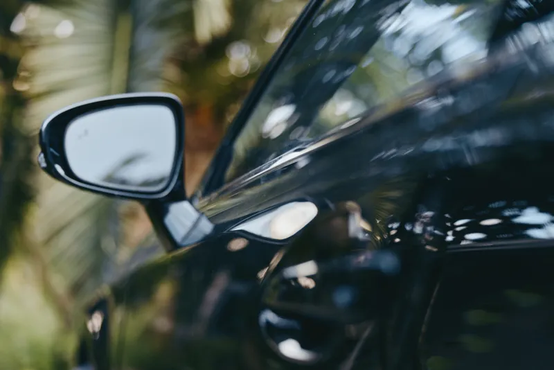 Car side mirror and door handle on glossy black car with tropical reflection, outdoor closeup. Sleek vehicle detail suggesting a mid30s driver in casual outfit, travel lifestyle and vacation vibe.