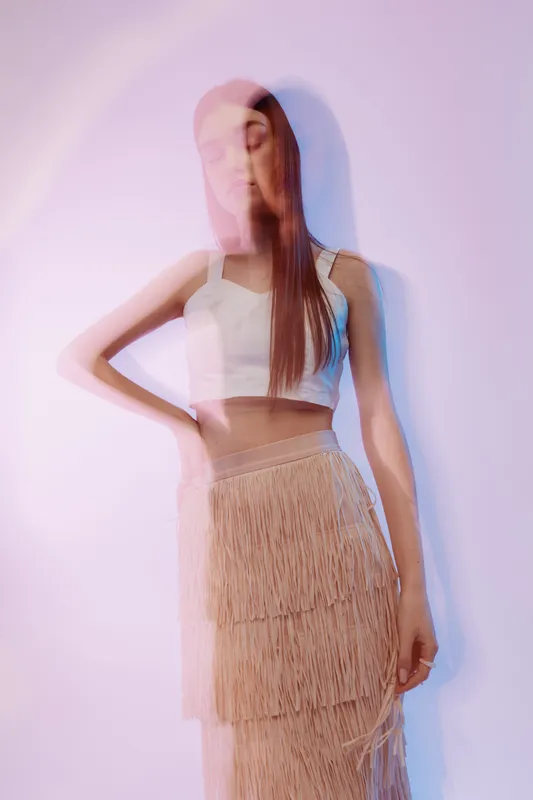 Young woman with long hair in a white crop top and fringed skirt poses against a light background with motion blur effect. Concept of beauty fashion and modern style.