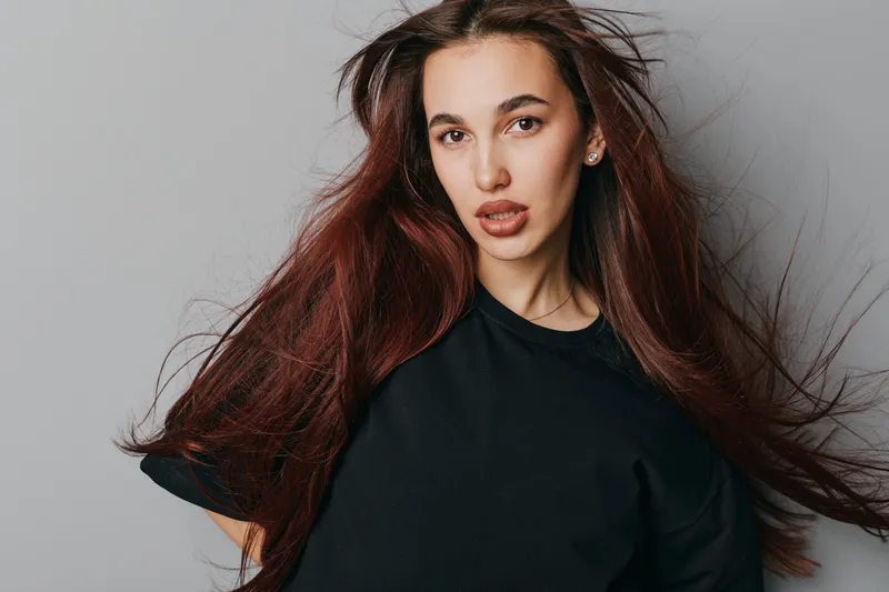 Young woman with long brown hair wearing black shirt posing indoors on gray background. Female model with natural makeup and confident expression in casual outfit for fashion portrait.