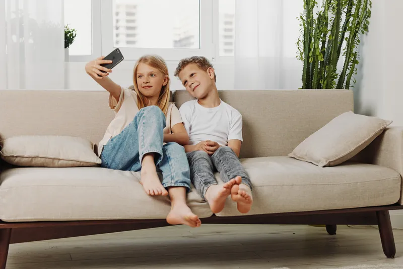 Two happy children sitting barefoot on beige sofa taking selfie with smartphone in bright living room. Casual clothes, cozy home atmosphere, family bonding and modern lifestyle concept.