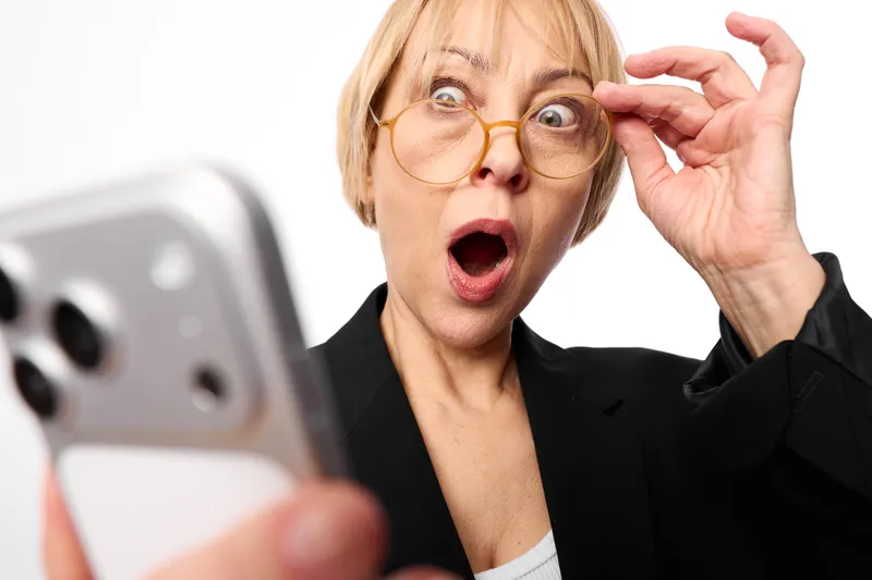 Surprised blonde woman with glasses in office, reaching with hand to her eyewear while reacting to a smartphone on desk, wide open mouth, intense emotion, bright modern light