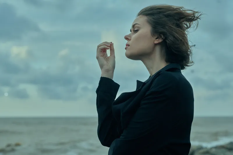 Woman in a dark blazer stands by the sea, profile view against a windy sky, conveying contemplative mood, fashion style and coastal storytelling for outdoor photography and editorial stock imagery