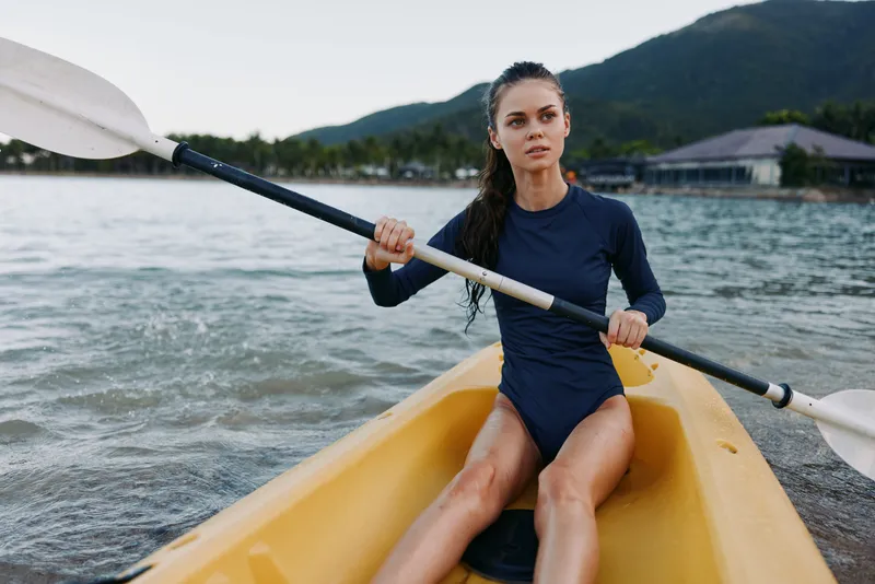 Female kayaker paddling a yellow kayak on calm coastal water, wearing a dark blue swimsuit, gripping a double-ended paddle with focus and determination.