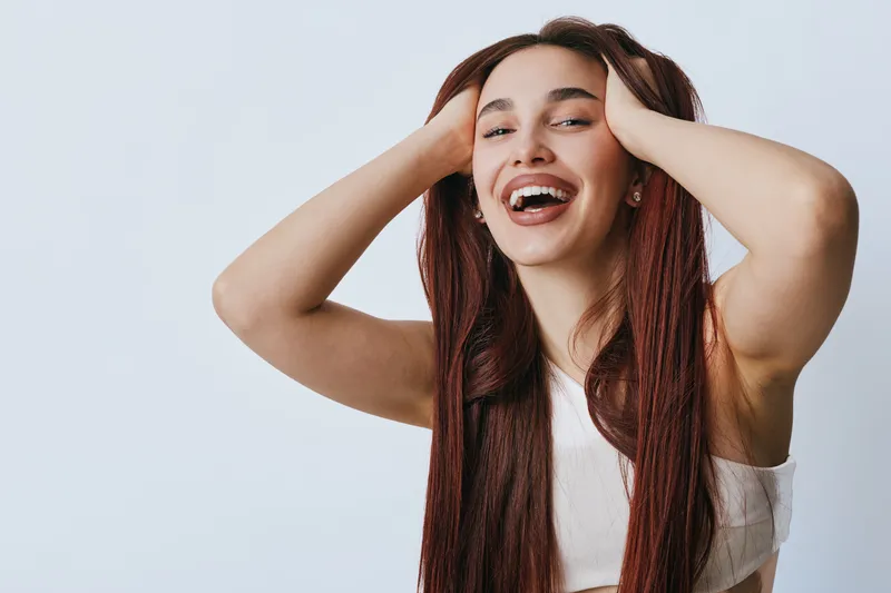 Young woman with long braided hair smiling and holding head with hands in casual sleeveless top against plain background. Happiness, joy and natural beauty with confident expression.