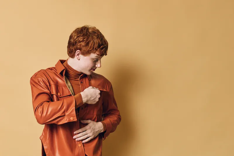 Redhead man with curly hair dressed in orange jacket and turtleneck shirt poses against beige background looking sideways with thoughtful expression and hand on chest.