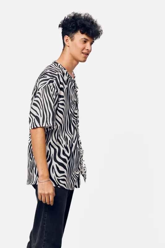 young man wearing casual zebra print shirt and black pants standing in profile against plain white background, confident pose with natural smile and curly dark hair