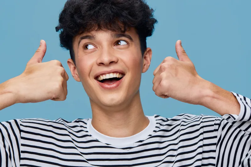 happy young man showing thumbs up gesture wearing striped shirt with cheerful expression on blue background, positive emotion and casual style