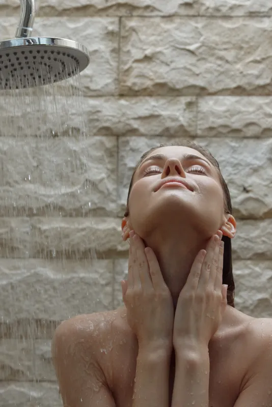 woman relaxing under shower with warm water pouring down, enjoying cleansing and refreshing moment with eyes closed against stone wall backdrop