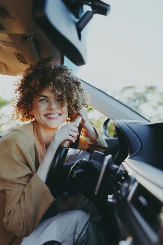 A smiling woman driver sits behind the wheel in a sunlit car interior, conveying a relaxed lifestyle moment with retro film color grading and warm tones, casual attire, candid pose, and everyday