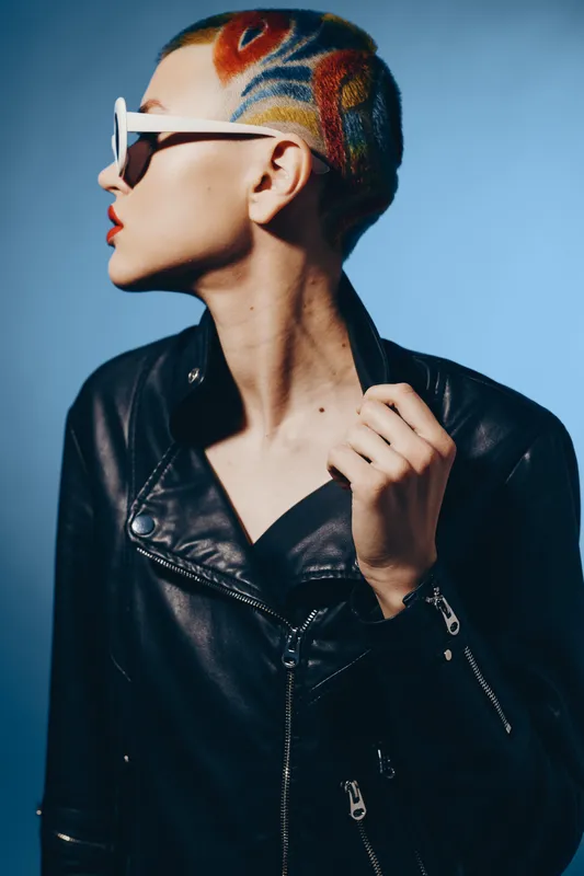 Young woman with colorful shaved hair and stylish sunglasses wearing a black leather jacket, posing confidently against a blue studio background in modern fashion style.