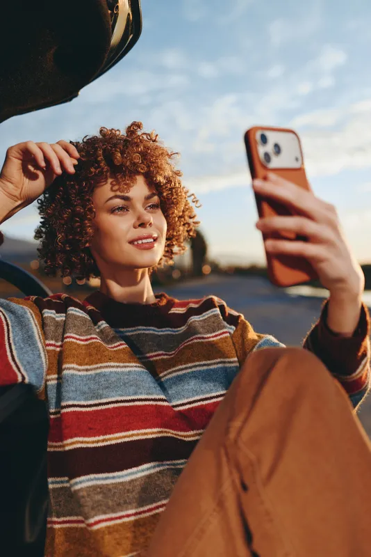 Woman lifestyle selfie moment, curly hair woman outdoors at sunset, holding smartphone and adjusting hat brim, relaxed expression, urban street background, warm evening light