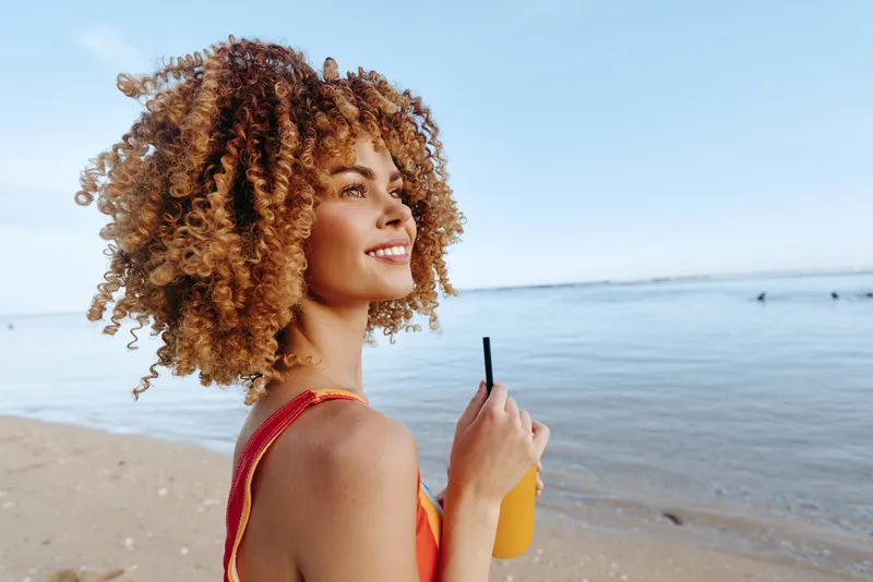 Woman with curly hair outdoors on sandy beach, holding sunglasses, side profile gaze to horizon, sunny summer afternoon, sea breeze, clear blue sky, calm ocean background, natural tones