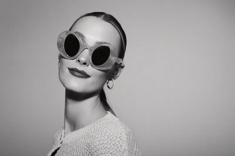 Stylish young white woman with pale skin wearing large round sunglasses and hoop earrings poses confidently in studio. Concept for fashion, accessories, and modern portrait.