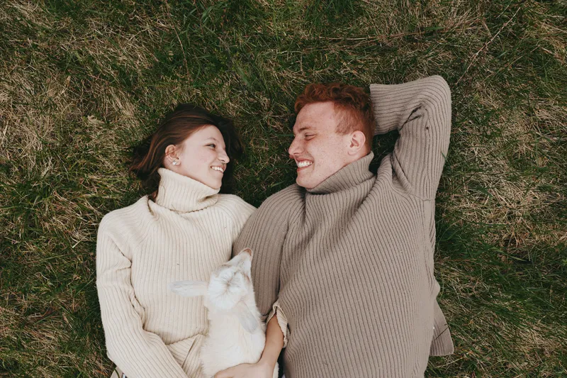 Smiling couple lying on grass in cozy sweaters, sharing a gentle moment with a goat kid nearby. Natural light, calm atmosphere, and lifestyle focus convey warmth, affection, and harmony with animals.