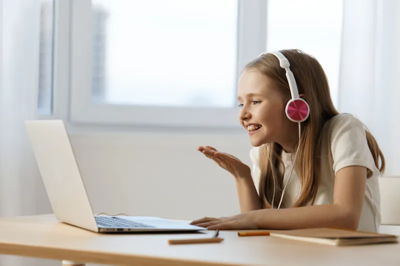 Happy girl with headphones engaging in an online class using a laptop, smiling and interacting, modern interior with natural light, educational atmosphere