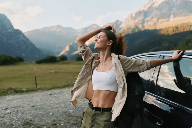 Young woman enjoying outdoor lifestyle beside a parked car in a valley under mountains, stretching arms, smiling, embracing freedom, nature, and adventure.