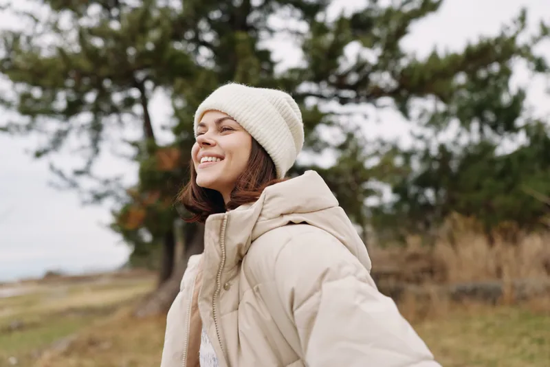 Woman in a beige coat and white beanie smiles in a park, trees and grass in the background, capturing a casual outdoor portrait with autumn light