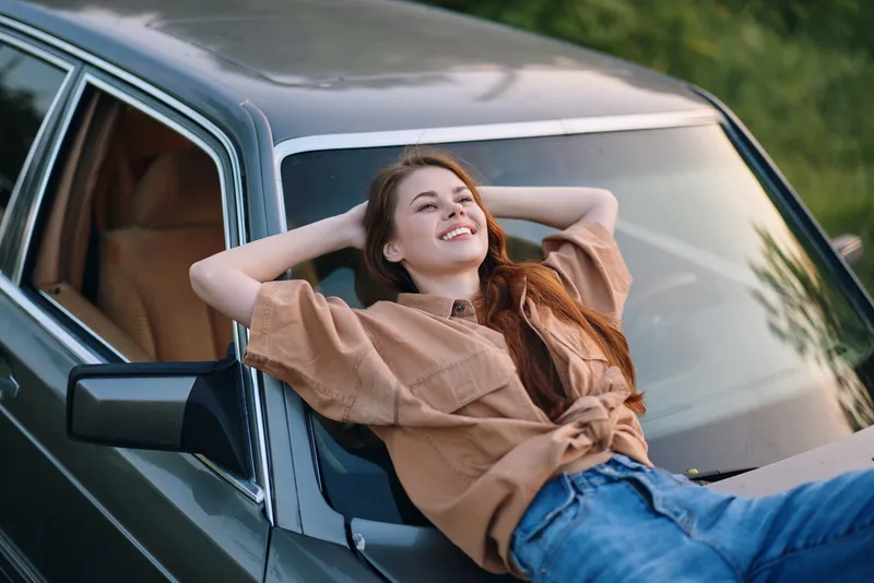 Happy young woman relaxing on a vintage car, enjoying the outdoor sun, dressed in casual brown shirt and denim jeans, surrounded by green nature.