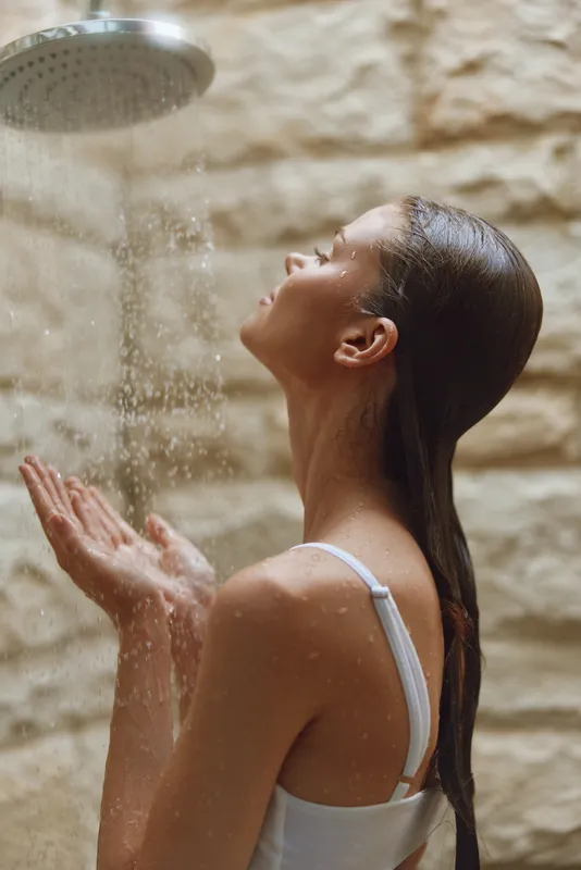 woman showering outdoors under rain-style showerhead with closed eyes, enjoying water droplets, wet hair, relaxing moment, beige textured wall background