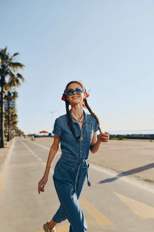 Smiling woman in denim jumpsuit enjoys sunny day outdoors with sunglasses, braided hair, and palm trees along a coastal promenade.
