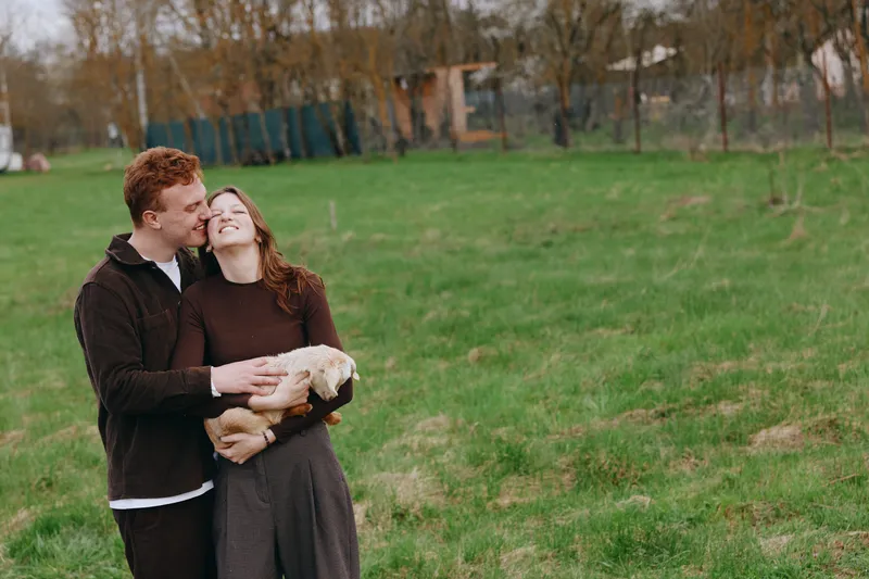 Cheerful couple outdoors with a goat kid, embracing and smiling in a sunlit field, rustic setting, natural colors, gentle breeze, relaxed mood, showing affection and companionship in a pastoral