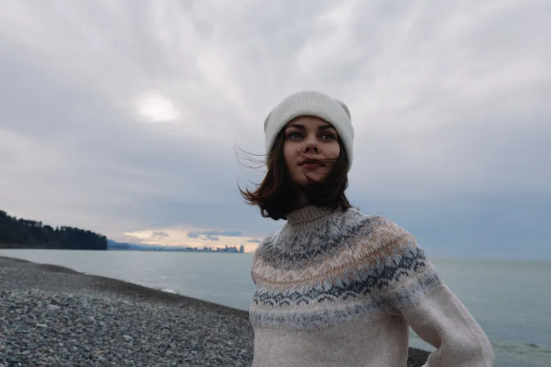 Woman on a windy beach wearing a knit sweater and beanie near the shoreline, gazing at the horizon with sea and cloudy sky as a chilly, relaxed outdoor portrait.