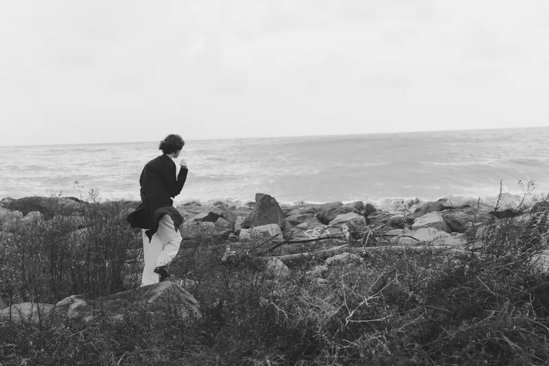 Coast scene with a distant sea, rugged rocks, and a lone silhouette walking along the shore in a monochrome landscape, capturing wind swept atmosphere and serene maritime wilderness