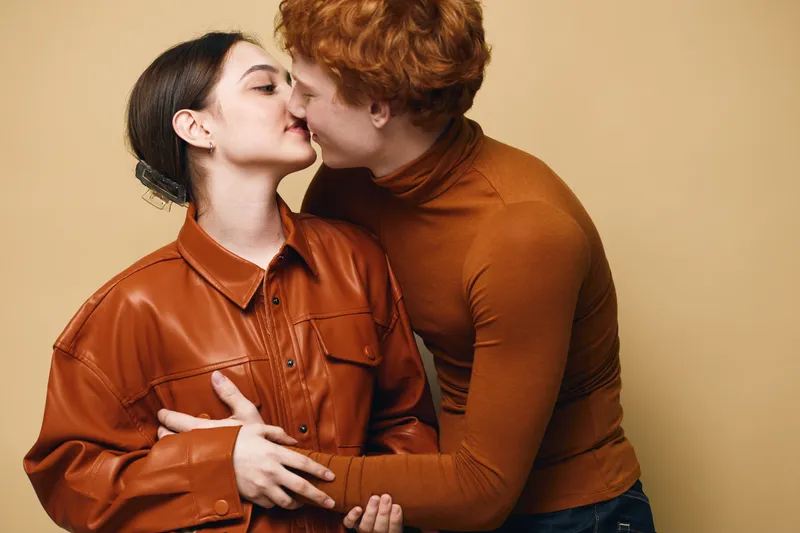 Couple, love, romance and affection with woman and man kissing and embracing against beige background. Intimacy, relationship, emotions and connection between young people in casual clothes.