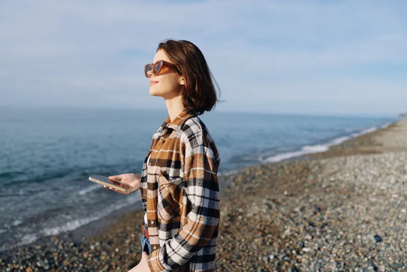 Woman on a pebbled beach with smartphone, wearing a plaid shirt and sunglasses, walking along the shoreline with calm sea and blue sky in the background, casual outdoor mood.