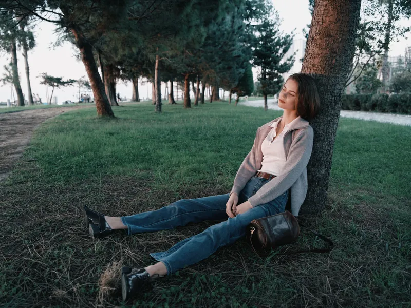 A thoughtful woman rests against a tree in a park, enjoying a quiet moment outdoors. Soft light, green grass, and peaceful surroundings create a calm, reflective atmosphere.